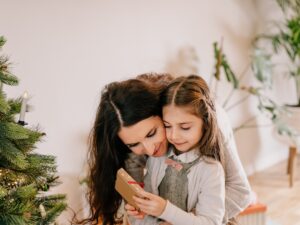 Smiling mother hugging her preschool-age daughter opening her present near Christmas tree