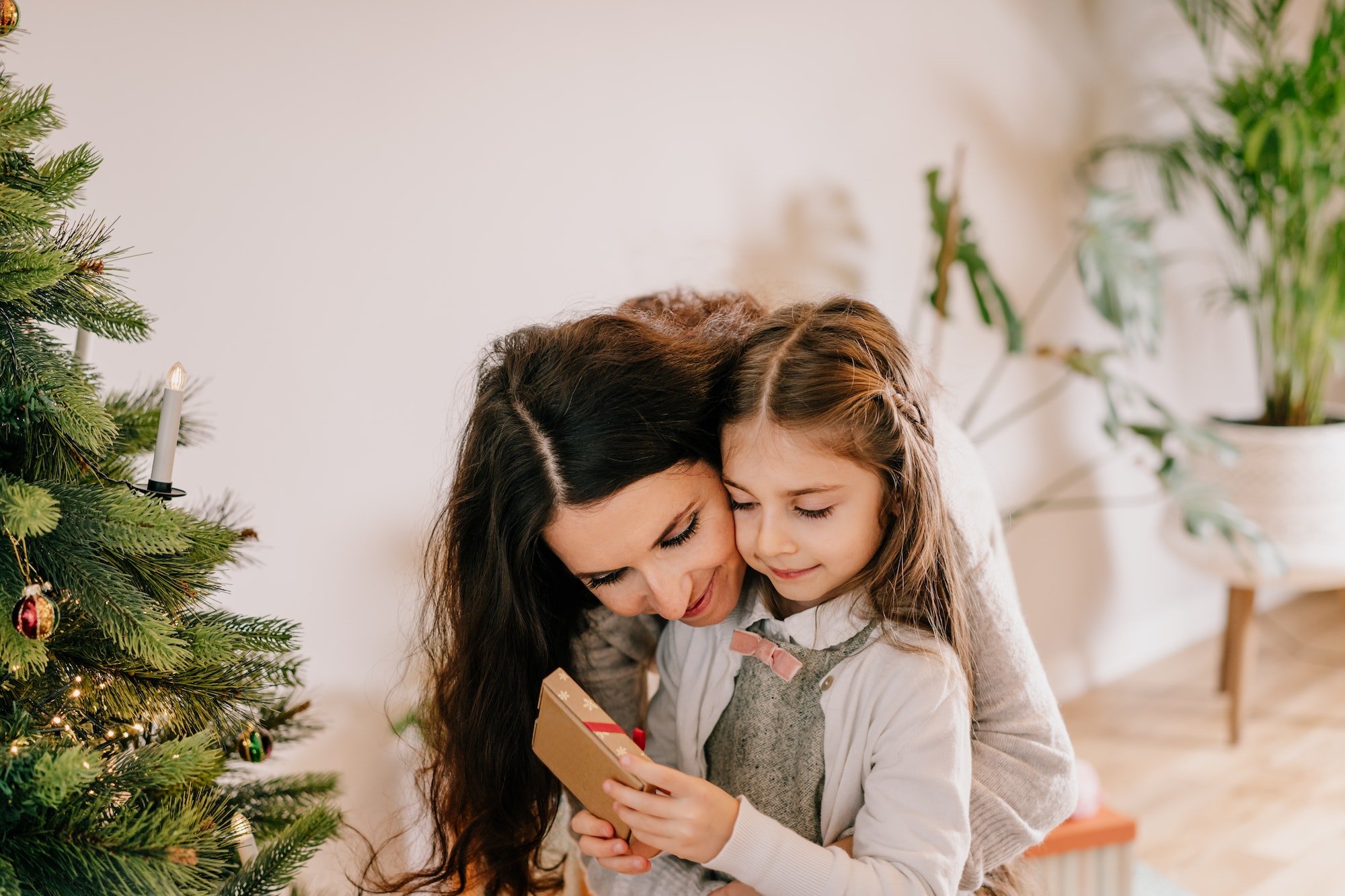 Smiling mother hugging her preschool-age daughter opening her present near Christmas tree