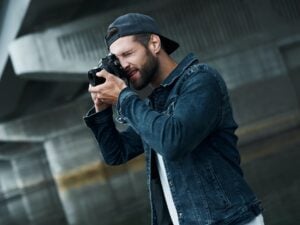 Photography hobby. Young stylish man standing on city street taking photos on camera concentrated